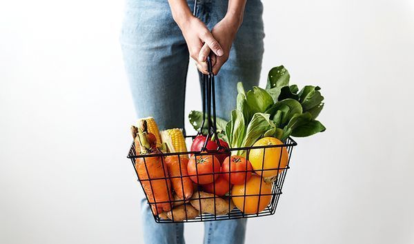 Nainen roikottaa vihanneksia ja hedelmiä sisältävää kauppakoria käsissään.